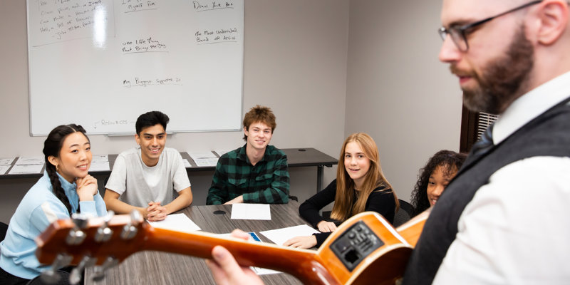 A group of smiling students engages in a music therapy session, led by an instructor with a guitar, fostering creativity and connection.