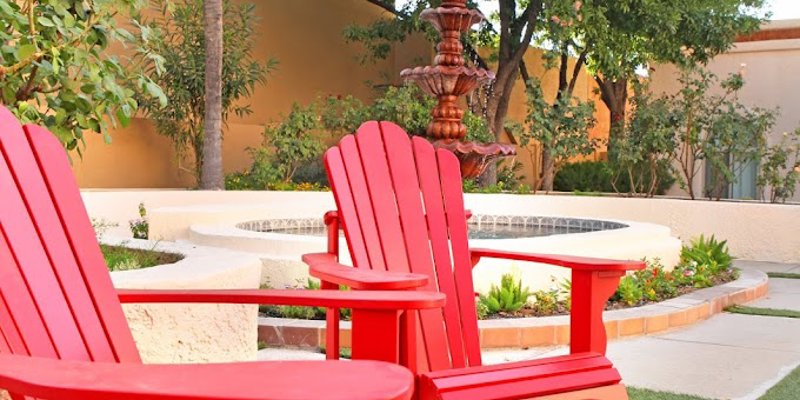 Vibrant red Adirondack chairs sit beside a tranquil fountain surrounded by lush greenery and palm trees, creating a peaceful outdoor retreat